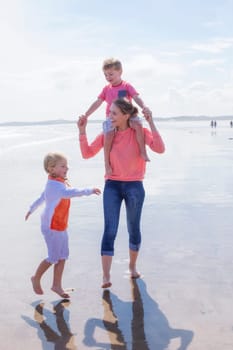 Happy single parent family walking along the beach. They are all laughing and smiling and walking across the waters edge. The daughter is walking alongside her mother while her son is on her shoulders.