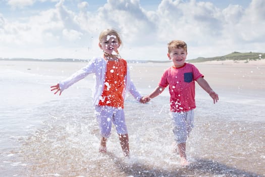 Brother and sister splashing in the water at the beach. They are wearing casual clothing and smiling.