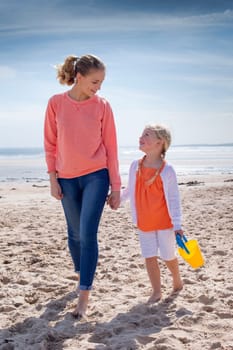 Young mother and daughter walking along with beach. They are looking at each other and smiling.