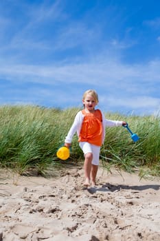 Little Girl running onto the beach with her arms outstretched and holding a bucket and spade.