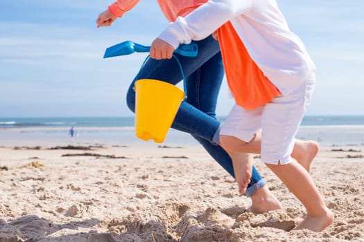 Mother and Daughter running along the beach. Only their legs can be seen, the little girl is holding a bucket and spade.