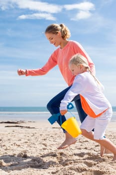 Young mother and daughter running along the beach with a bucket and spade. They are both laughing and smiling.