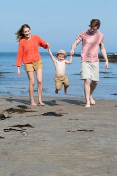 Young two parent family at the beach with their young son. They are all holding hands and swinging the little boy.