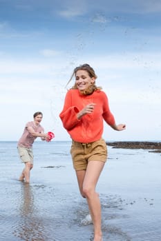 Portrait of a beautiful young couple at the beach. He is holding a bucket and splashing her as she runs away from him.