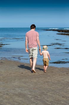 Father and son bonding on the beach. They are wearing casual clothing and walking toward the sea edge holding hands.