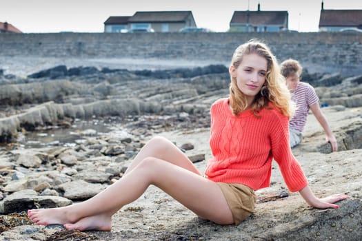 Young girl sitting on a rock at the beach wearing casual clothing. Her boyfriend can be seen in the background.