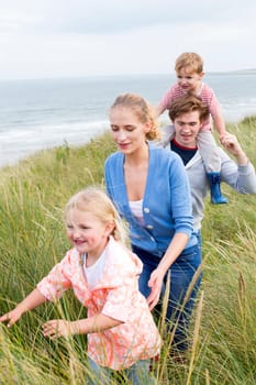 Close up of a young family of four walking through long grass next to the beach. They are wearing casual clothing and are smiling.