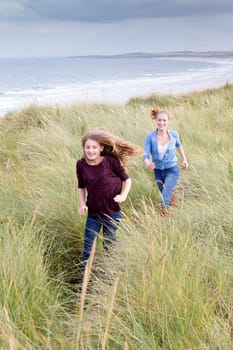 Two sisters running through long grass with the background of the beach. They are wearing casual clothing and smiling.