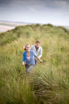 Young couple running through long grass with the background of the beach. They are both smiling and wearing casual clothing.