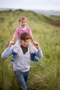 Portrait of a father and son walking through long grass next to a beach. The little boy is on his fathers shoulders and they are both smiling.
