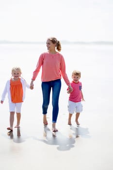 Young mother and her two children walking along the beach. They are barefoot and wearing casual clothing.