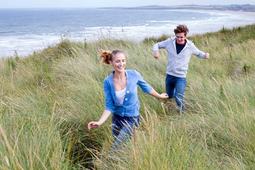 Young couple running through long grass with the background of the beach. They are both smiling and wearing casual clothing.