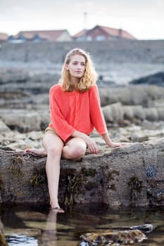 Beautiful young woman sitting on a rock at the beach. She is barefoot and is wearing casual clothing.
