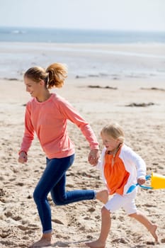 Young mother and daughter running along the beach with a bucket and spade. They are both laughing and smiling.