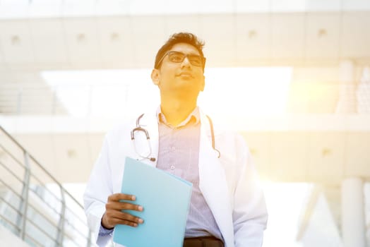Asian Indian medical doctor standing outside hospital building, beautiful golden sunlight at background.