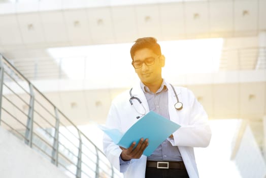 Portrait of Asian Indian medical doctor reading on medical report, standing outside hospital building, beautiful golden sunlight at background.
