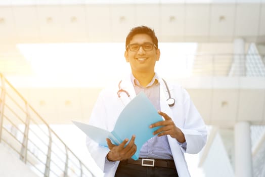 Portrait of Asian Indian medical doctor smiling and reading on medical report, standing outside hospital building, beautiful golden sunlight at background.