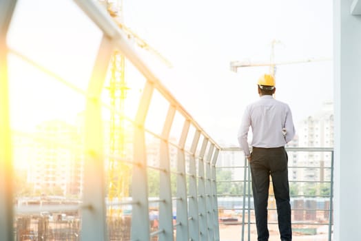 Rear view Asian Indian male site contractor engineer with hard hat holding blue print paper inspecting at construction site, crane with golden sunlight at the background.