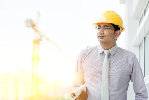 Handsome Asian Indian male site contractor engineer with hard hat holding blue print paper walking at construction site, crane with golden sunlight at the background.