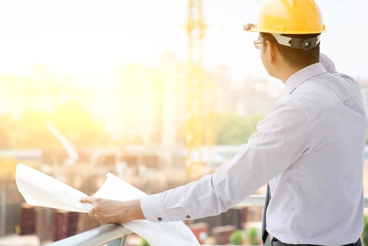 Asian Indian male site contractor engineer with hard hat holding blue print paper looking away inspecting progress at construction site, crane with golden sunlight at the background.