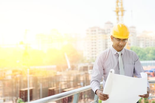 Asian Indian male site contractor engineer with hard hat holding blue print paper inspecting at construction site, crane with golden sunlight at the background.