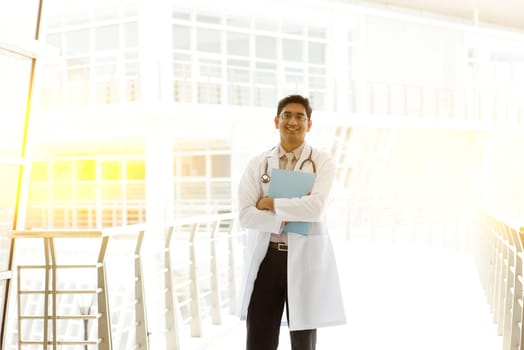 Portrait of Asian Indian medical doctor holding medical report, standing outside hospital building block, beautiful golden sunlight at background.