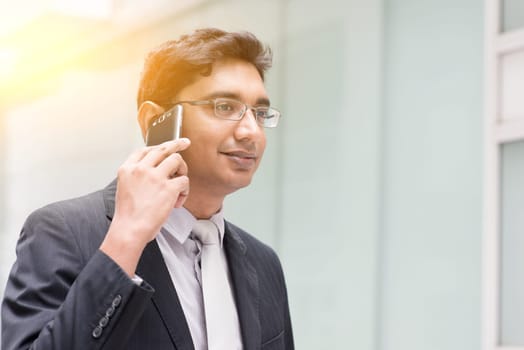 Portrait of Asian Indian business man using smartphone, outside modern office building block, beautiful golden sunlight at background.