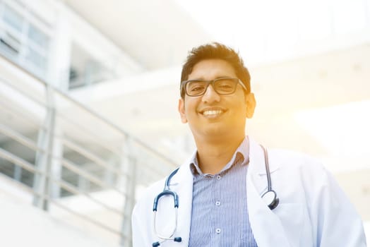 Portrait of Asian Indian medical doctor smiling and looking at camera, standing outside hospital building, beautiful golden sunlight at background.