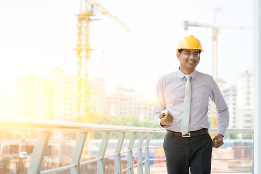 Asian Indian male site contractor engineer with hard hat holding blue print paper walking at construction site, crane with golden sunlight at the background.