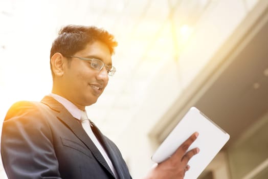 Portrait of Asian Indian business man using touch screen tablet computer, outside modern office building block, beautiful golden sunlight at background.