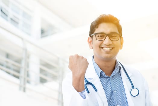 Portrait of successful Asian Indian medical doctor smiling and celebrating success, standing outside hospital building, beautiful golden sunlight at background.