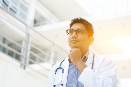 Portrait of Asian Indian medical doctor thinking and looking away, standing outside hospital building, beautiful golden sunlight at background.
