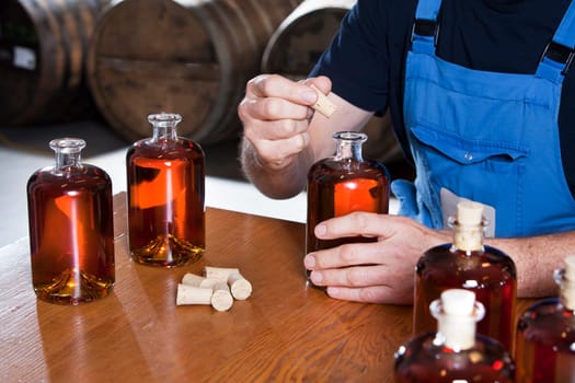 Putting the cork on the bottle of just bottled liquor and some barrels in the background