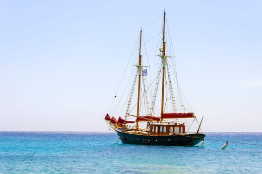 A traditional wooden sail boat anchored on the coast of Mykonos, Greece. The wind sails are down and it is tied on the rocks by a rope. A very typical greek island summer holiday scene on crystal clear blue sea.