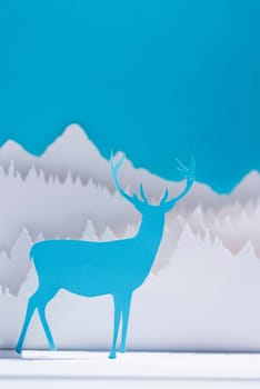 Holiday paper cut blue reindeer in winter forest background. Handmade template ideal for Christmas greeting card, New Year or seasonal campaign.