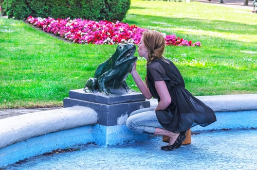 Young woman kisses the frog sculpture in a fountain.