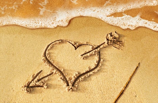 Heart with arrow, as love sign, drawn on the beach shore, with some water and foam coming from the sea.