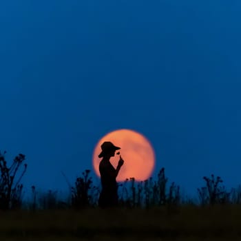 A woman with a hat in front of a full moon smells of a flower