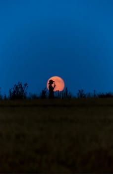 A woman with a hat in front of a full moon smells of a flower