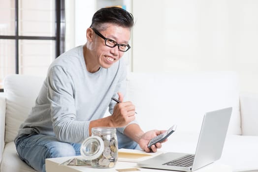 Mature 50s Asian man giving thumb up while counting on money using calculator and laptop computer. Saving, retirement, retirees financial planning concept.