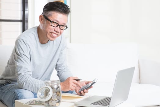 Mature 50s Asian man counting on money using calculator and laptop computer. Saving, retirement, retirees financial planning concept.