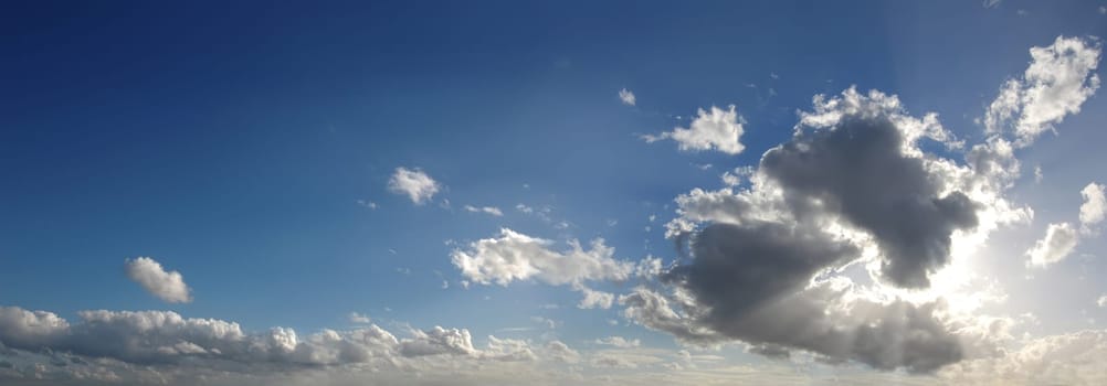 Blue sky with several cumulus clouds in background and a large cloud masking the sun, in the foreground, generating some godrays. Cinemascope format.