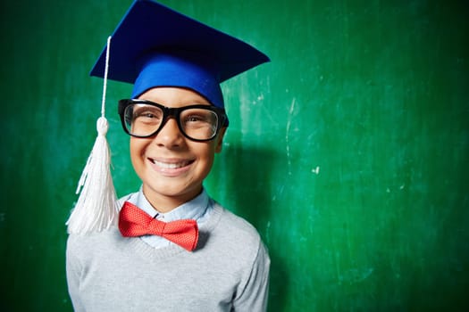 Joyful kid in eyeglasses and graduation hat looking at camera with smile