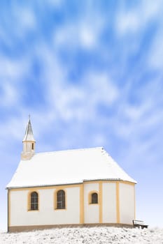 Tiny winter church or single snowbound small chapel vertical stock photo with blured in motion clouds in blue sky