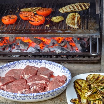 Grilling vegetables on barbecue, tomatoes, zucchini and eggplants, with a dish of beef meat waiting to be cooked