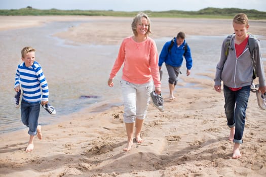 Family of four walking through sand barefoot at the beach. They are wearing warm casual clothing and are looking at the surroundings.