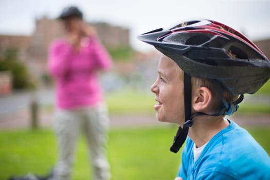 Child in the foreground with a bicycle helmet on while his mother is in the background taking a picture of him.