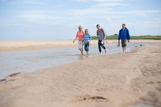 Family of four walking through water and sand at the beach. They are barefoot and wearing warm and casual clothing.