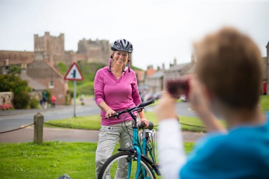 Mother posing for a picture on her bike. A village with a castle can be seen in the background, she is wearing a helmet and casual clothing.