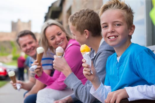 Family enjoying ice cream outside of a shop in a village. They are all holding their cones and a castle can be seen in the background in the distance.